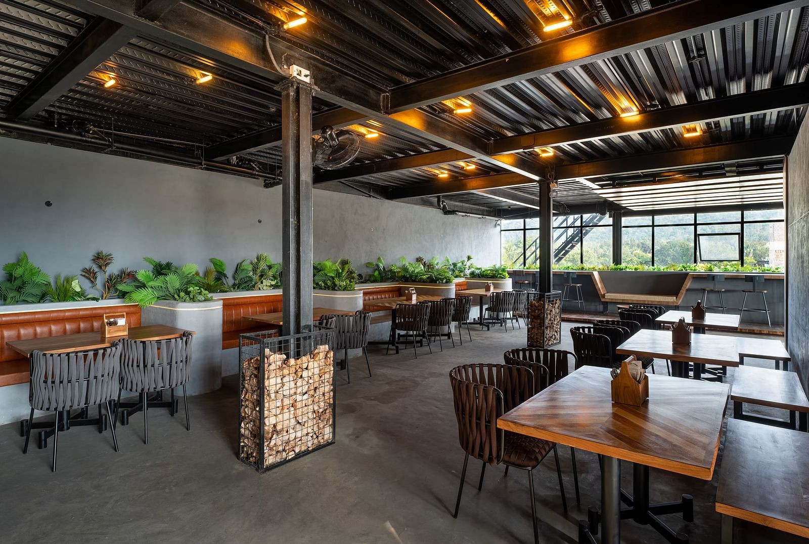 A modern open-air restaurant with wooden tables, cushioned seating, industrial-style ceiling, and lush greenery, offering a stylish dining experience at TGI Livancia Hillside, JC Nagar, Mysore.