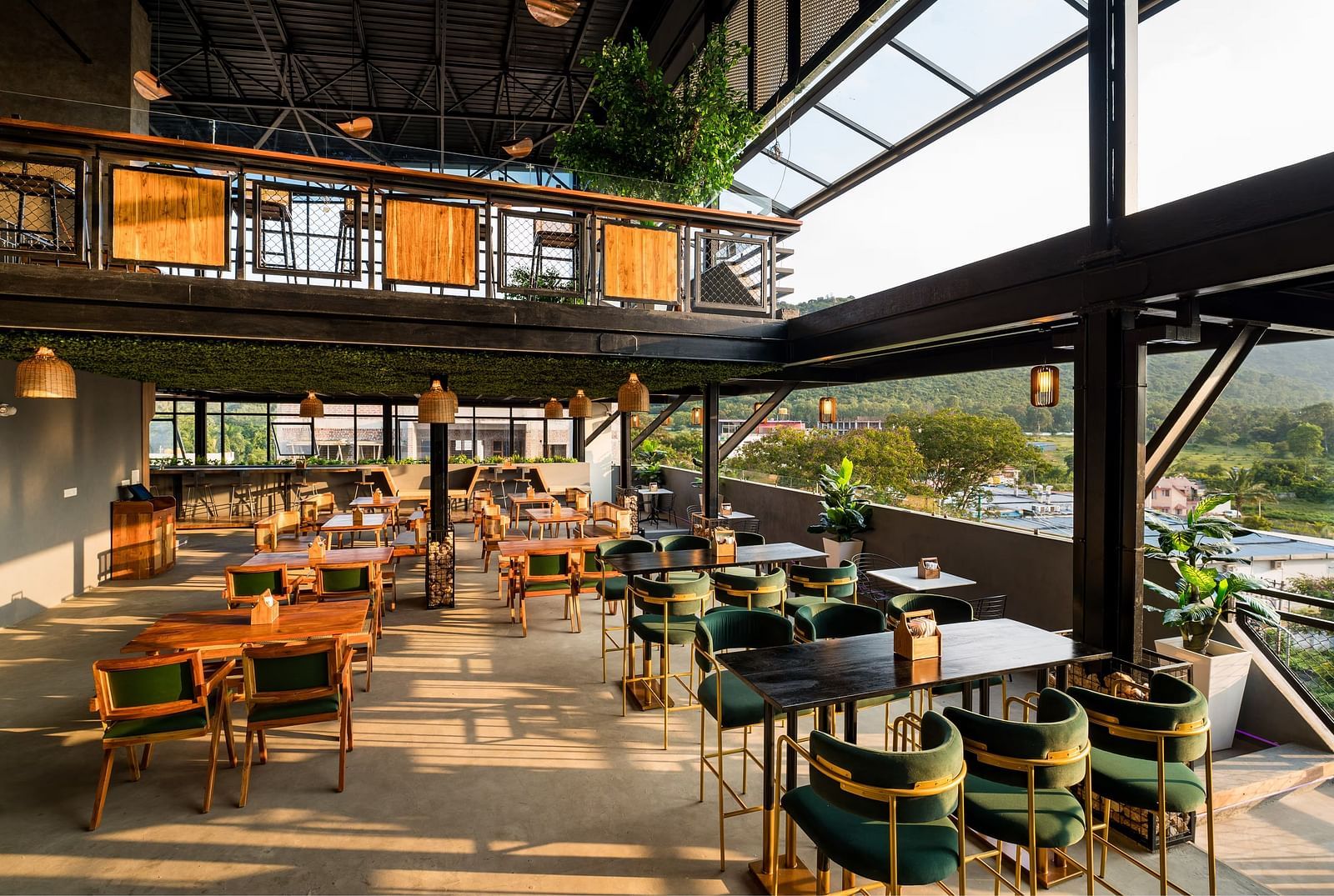 A spacious rooftop restaurant with elegant wooden furnishings, green velvet chairs, and panoramic views of the hills, bathed in warm natural light at TGI Livancia Hillside, JC Nagar, Mysore.