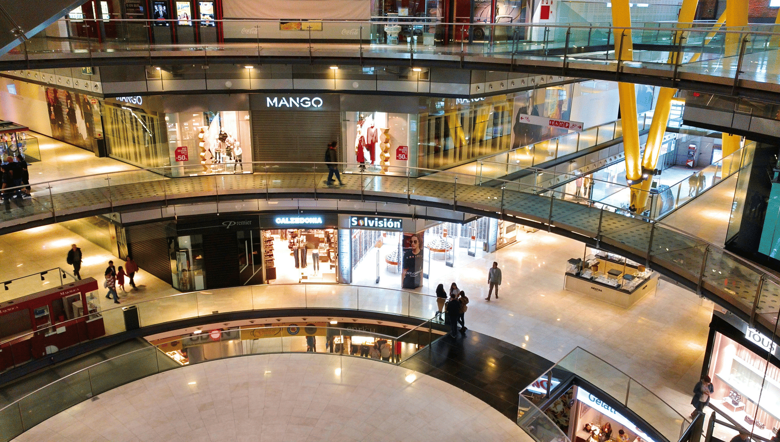Interior view of a modern shopping mall with multiple floors, glass railings, and people walking around the central atrium