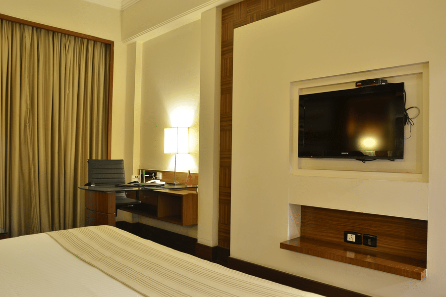 Premium Room at The Bristol Hotel, Gurgaon with a work desk and black chair, a desk lamp, a wall-mounted flat-screen TV in a wood-trimmed niche, and curtains.