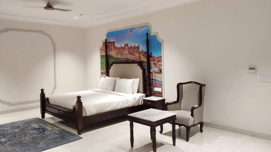 A bedroom at Anant Mahal, Jaipur featuring a king size bed, a small wooden table with a chair, a decorative mural on the wall, a ceiling fan and an A/C.