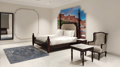 A bedroom view at Anant Mahal, Jaipur featuring a king size bed, a blue rug, a mirror, a seating area with a small table and a cushioned chair.