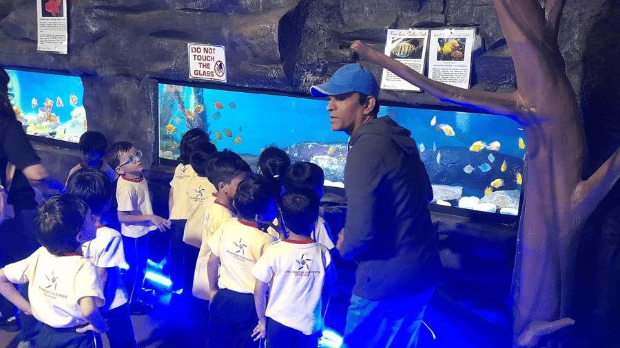 Visitors at EsselWorld Fish Paradise, including children and adults, stand in a dimly lit aquarium room, looking at various large fish tanks with bright blue and purple lighting.ighting.