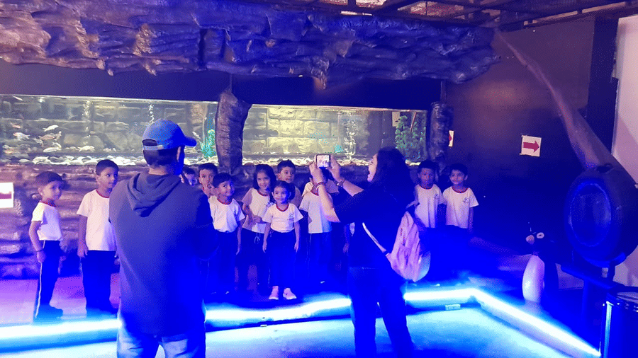 Visitors at EsselWorld Fish Paradise are gathered in a dark exhibition area, illuminated by bright blue and purple lighting emanating from the surrounding fish tanks.