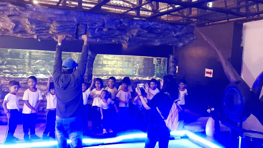 Visitors at EsselWorld Fish Paradise, including children and adults, stand in a dimly lit aquarium room, looking at various large fish tanks with bright blue and purple lighting.