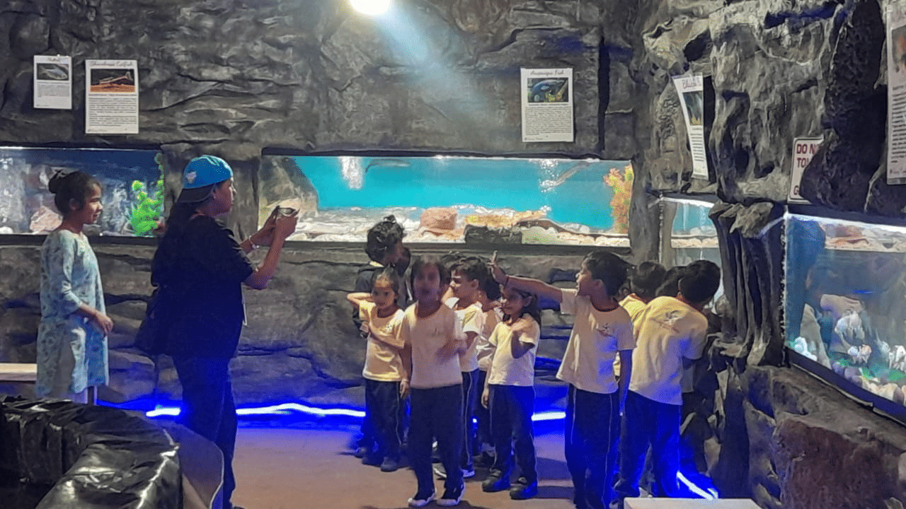 Students on a field trip at EsselWorld Fish Paradise stand in a wide-view aquarium room, looking at several large fish tanks lining the walls.
