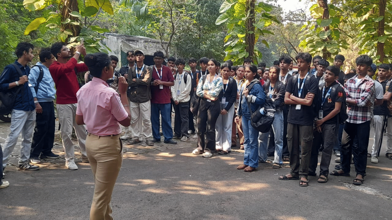 A group of students from EsselWorld Fish Paradise, possibly on a field trip, stand outdoors in a sunny park setting, listening to an adult guide or teacher.