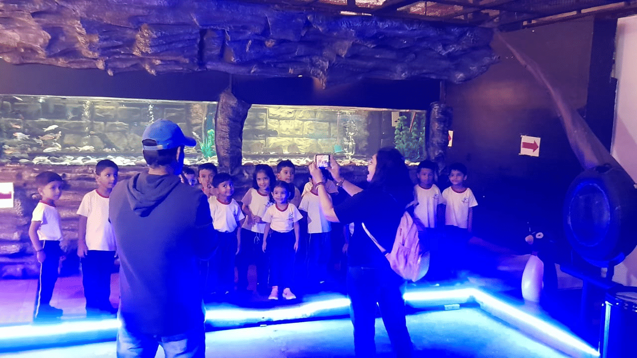 Visitors at EsselWorld Fish Paradise are gathered in a dark exhibition area, illuminated by bright blue and purple lighting emanating from the surrounding fish tanks.