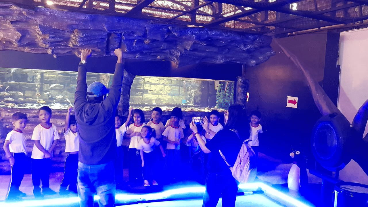 Visitors at EsselWorld Fish Paradise, including children and adults, stand in a dimly lit aquarium room, looking at various large fish tanks with bright blue and purple lighting.