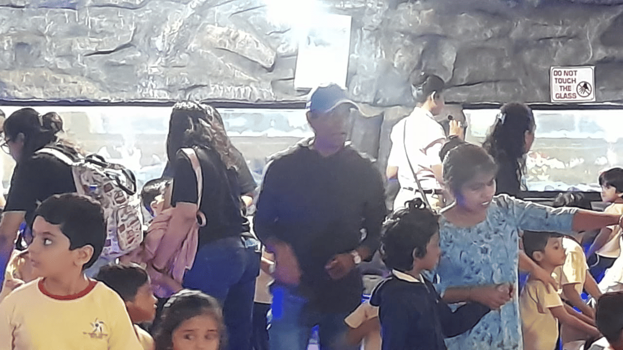 A group of children at EsselWorld Fish Paradise stand near a large fish tank, with adults in the background supervising the visit.