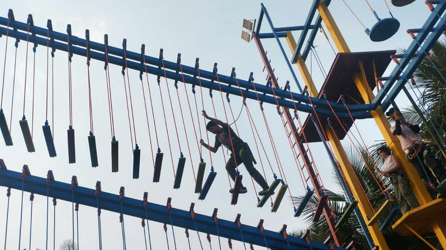 A person climbing a vertical wall with rope and ladder at an outdoor adventure activity zone at Vedic Village Spa Resort surrounded by lush tropical greenery.