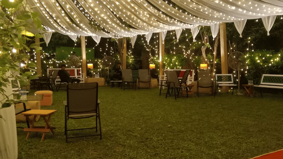 An outdoor dining setup at Vedic Village Spa Resort featuring tables and chairs arranged on a green lawn under a canopy of warm string lights in an evening setting.