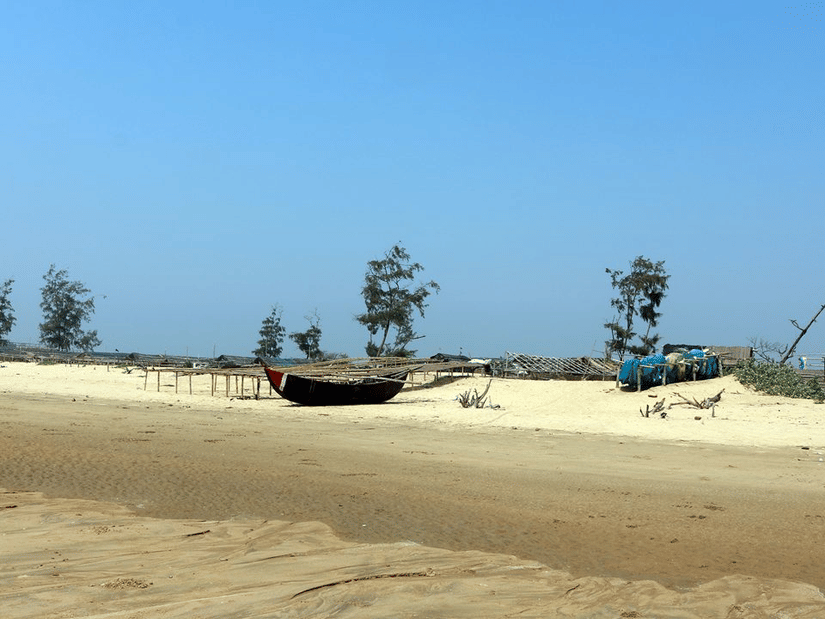 A traditional wooden fishing boat rests on the quiet, sandy shores of Shankarpur Beach near scattered fishing gear and coastal trees.