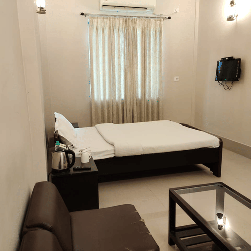 A room at Lake Side Resort Neermahal, featuring a single bed, bedspreads, a pillows, a sofa, a table, a window covered with curtains, and wall-mounted lamps.