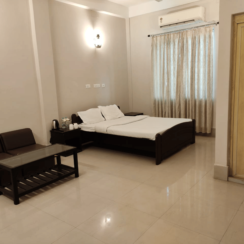 A room at Lake Side Resort Neermahal, featuring a large bed, bedspreads, pillows, a sofa, a table and a window covered with curtains.