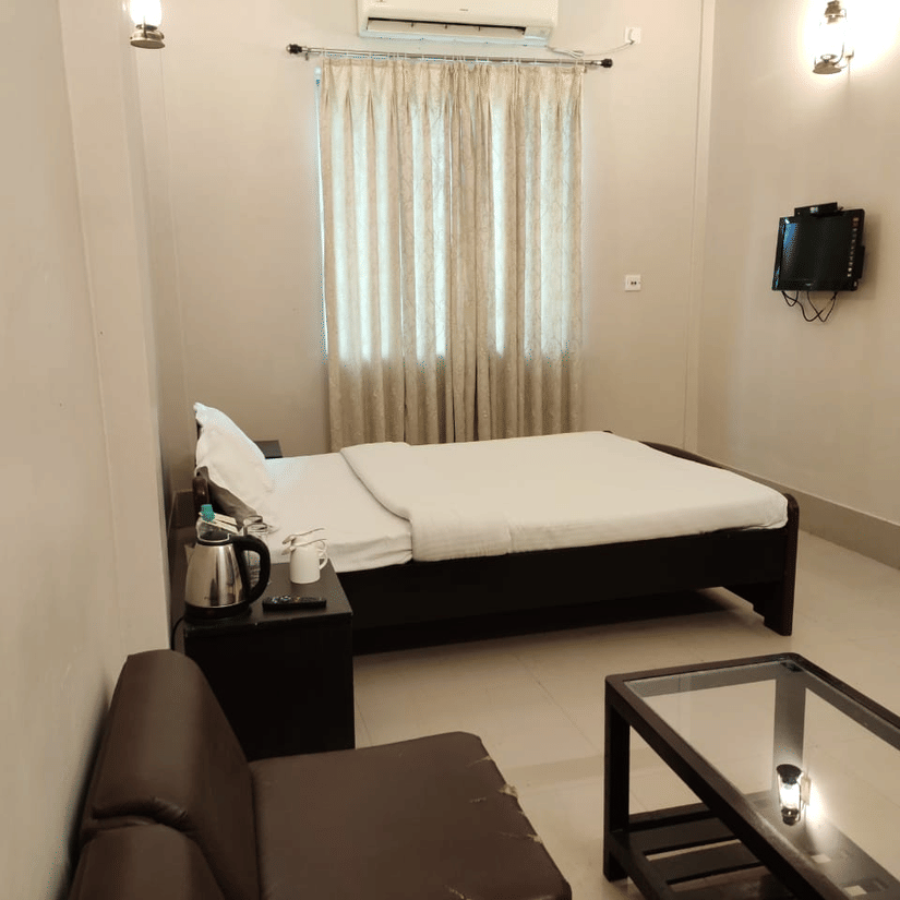 A room at Lake Side Resort Neermahal, featuring a single bed, bedspreads, a pillows, a sofa, a table, a window covered with curtains, and wall-mounted lamps.