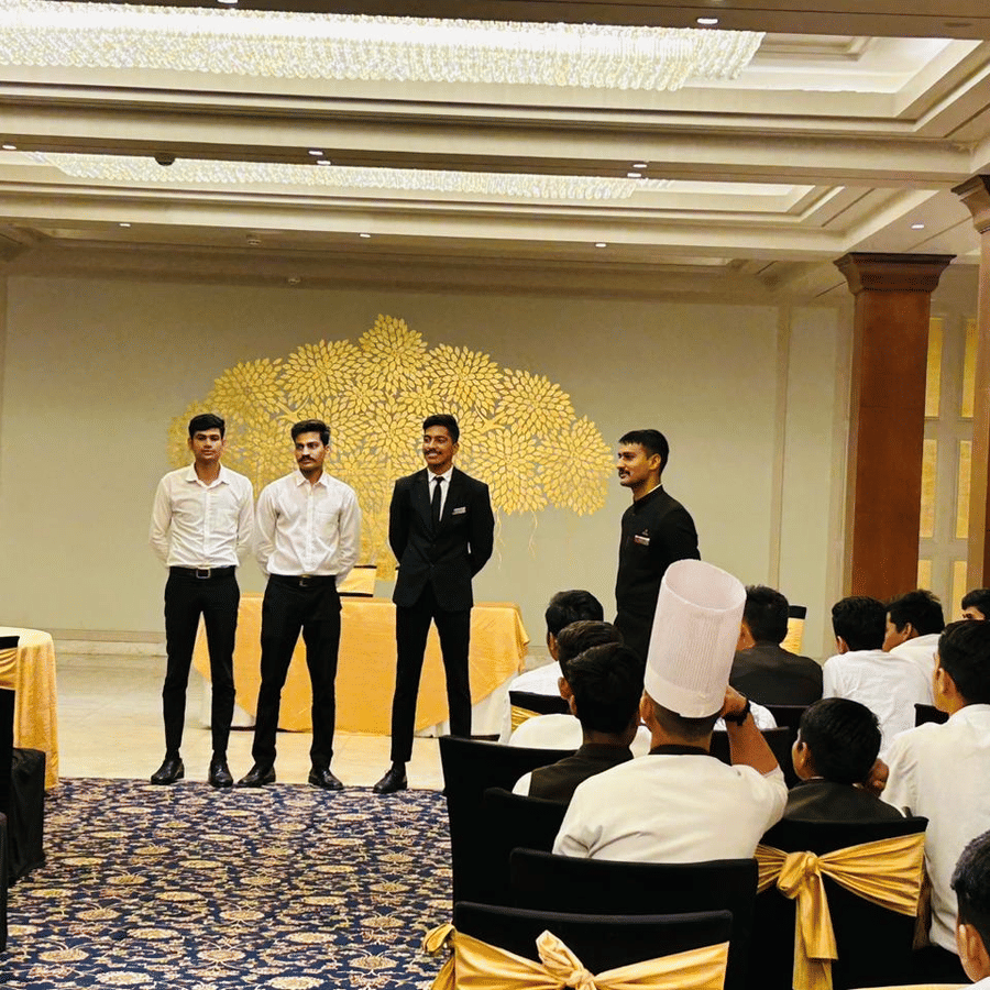 A line of individuals stands facing an audience during a gathering inside Clarks Group of Hotels. People sit at tables.