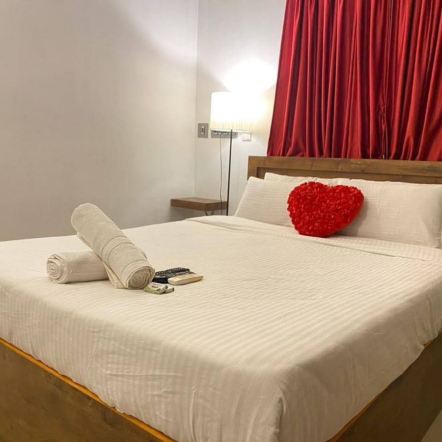alt-text A bed, with bedding and a heart shaped cushion, with a side lamp beside the bed in a bedroom | La Paradise Residency, Pondicherry
