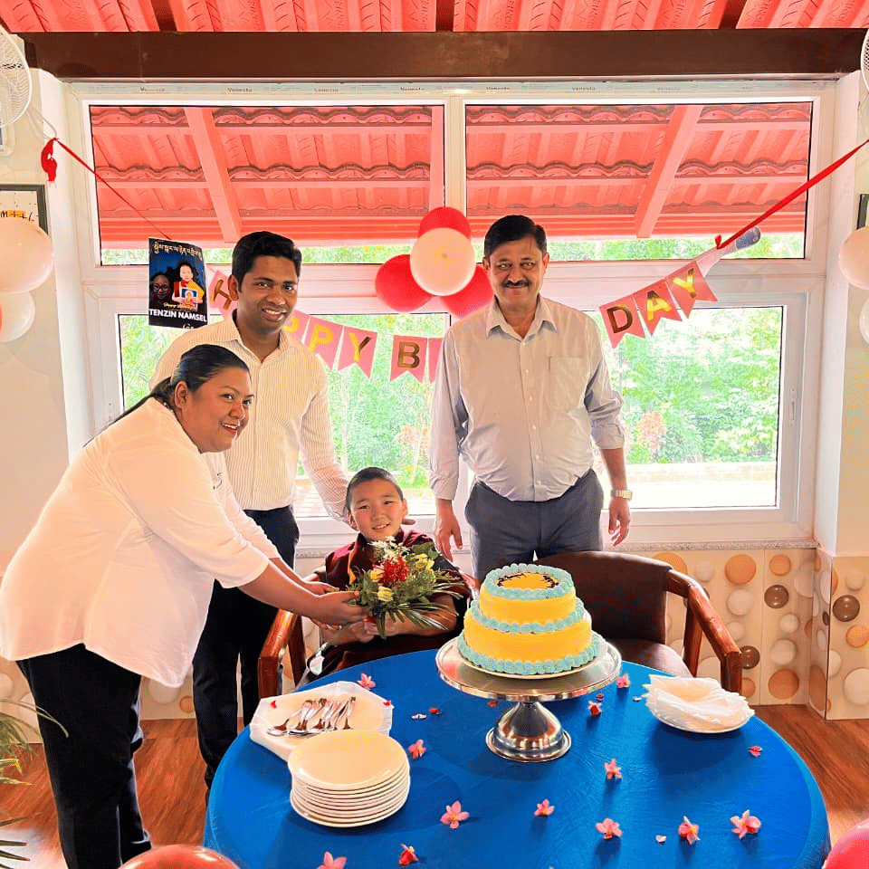 A family celebrating a birthday indoors, standing around a blue table with a yellow cake and balloons, with a red-tiled roof visible at Coorg Orange Blossom Resort and Spa.