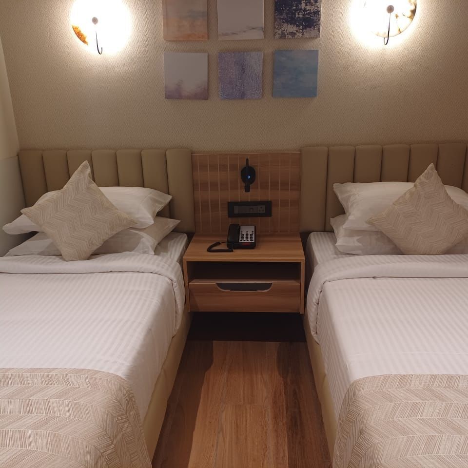Premium Room at Hotel Paai, Hosur featuring twin beds placed at two corners of the room. Above each bed there are lights on the wall. Between the beds there is a bedside table with a telephone and socket. 