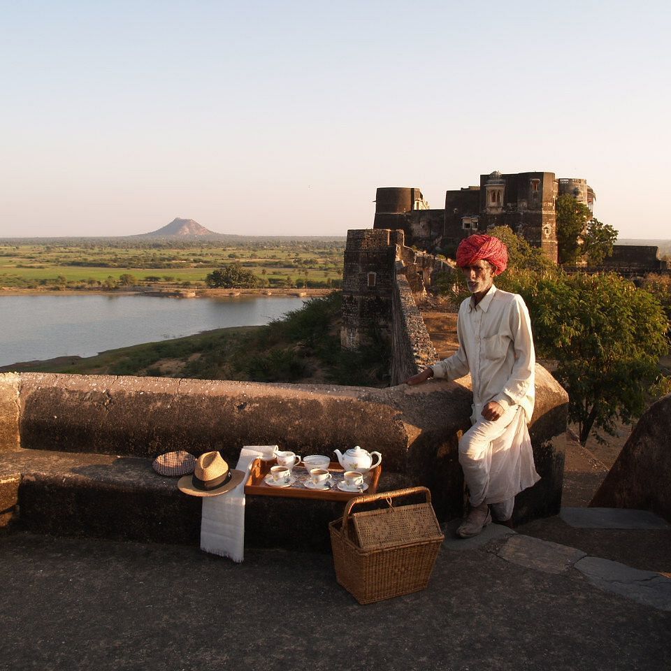 A man in traditional attire sits on a stone wall overlooking a landscape with a body of water, with a tea set laid out on a table beside him- Shahpura Bagh.