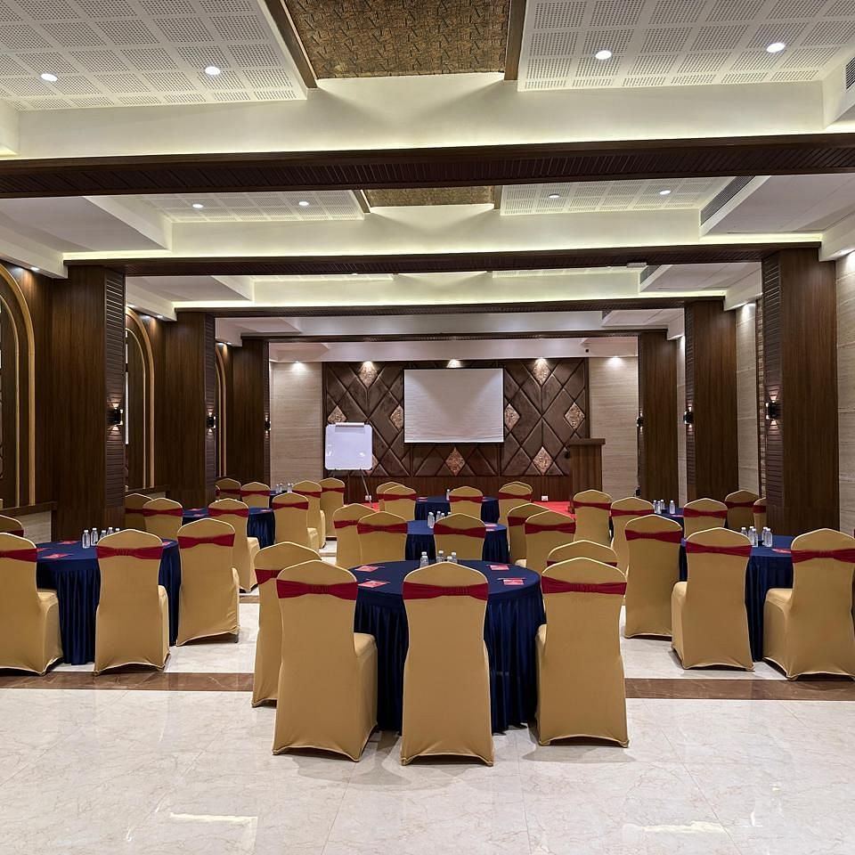 A view of a spacious contemporary hall featuring multiple decorated round tables and chairs around it facing a mounted screen at the backdrop at Shyanu Lords Eco Inn Airoli.