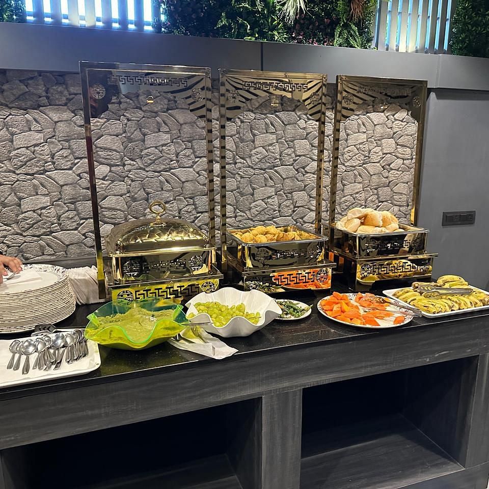 A buffet spread on a table set against a brick wall showcasing different food and fruits with a partially visible staff on the left at Shyanu Lords Eco Inn Airoli.