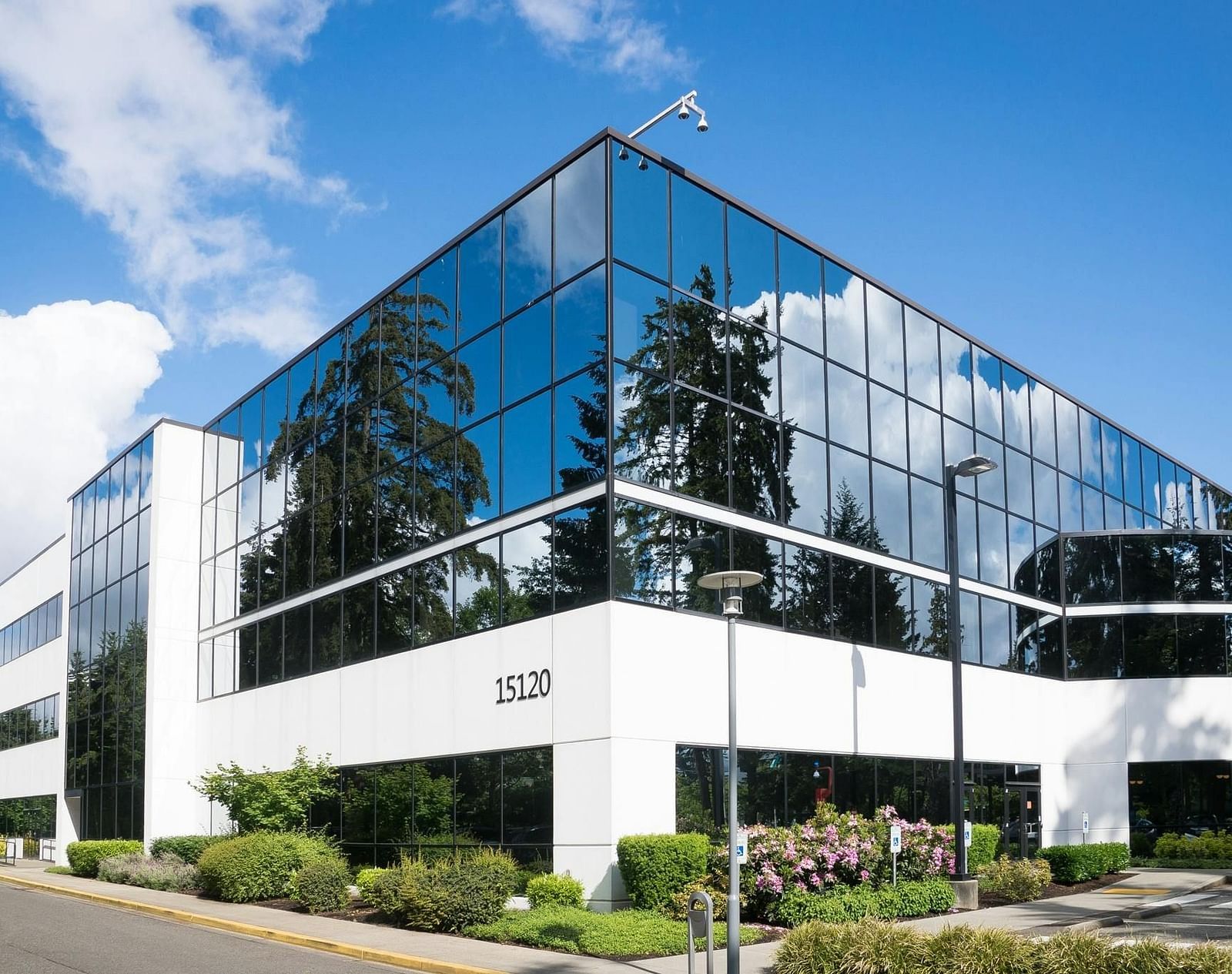 A modern, angular office building with glass walls reflecting the blue sky and surrounding greenery.