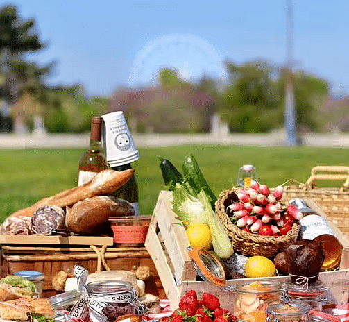 A rustic outdoor picnic with bread, wine, cheeses, fruits, and vegetables arranged on a red and white checkered blanket