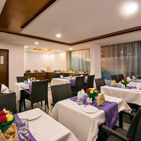 A dining area with tables covered in white cloths, place settings, a flower centrepiece and modern decor and lighting at at Lords Eco Inn Jayanagar Bangalore.