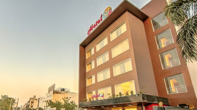 An exterior view of Hotel 91 HUDA Premier, Gurugram building with signage, entrance, parked cars, and surrounding road.