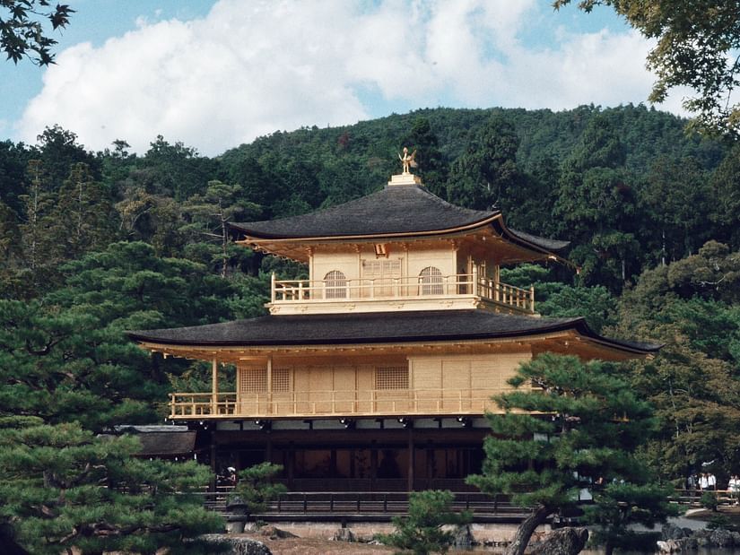 An image of the front view of a temple surrounded by lush green forest and the backdrop of a mountain