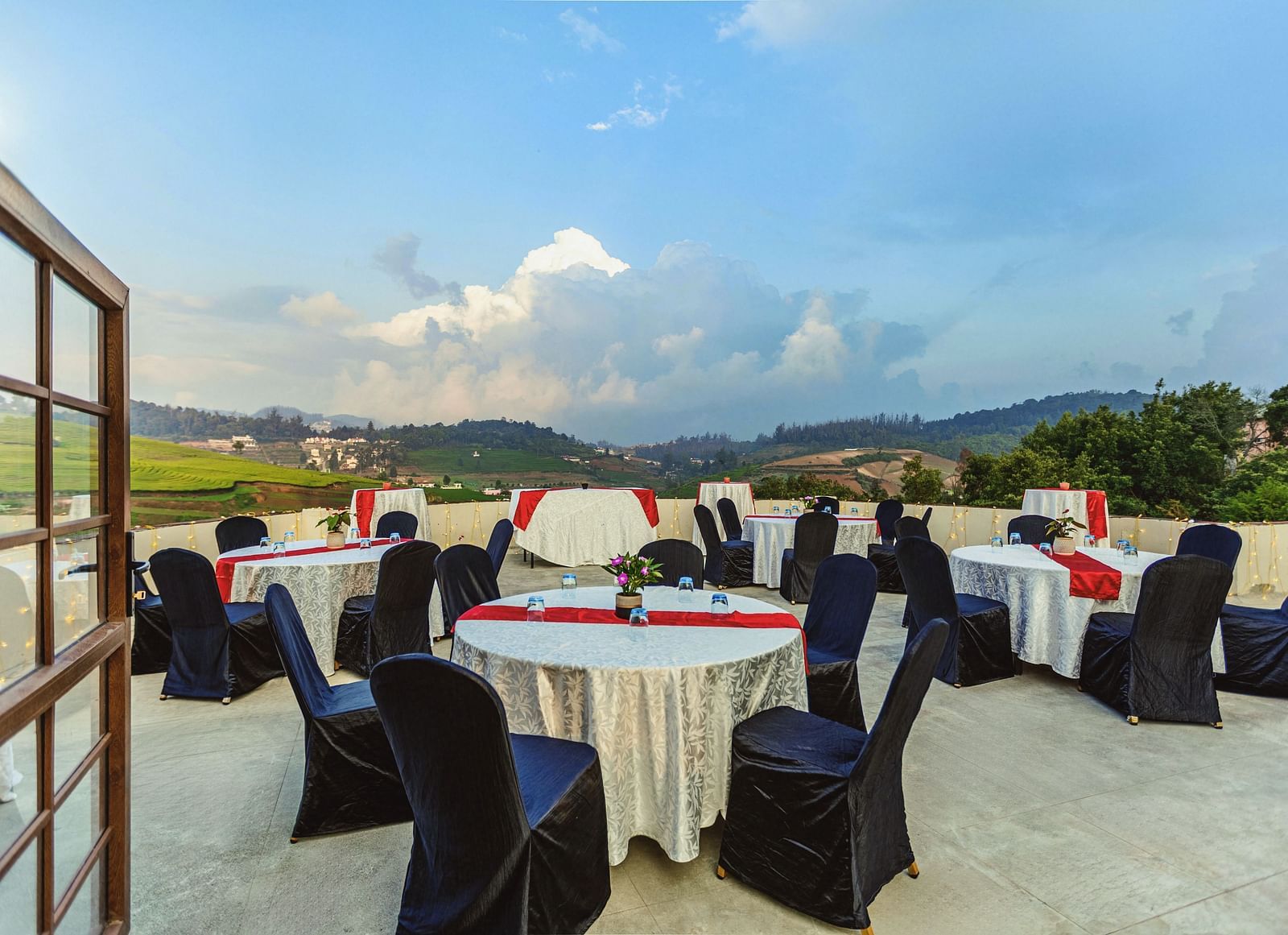 Rooftop event setup at Mango Hill Shola, Ooty, featuring round tables covered in white linens with red and black accents, surrounded by black banquet chairs, overlooking lush green hills and a dramatic cloudy sky.
