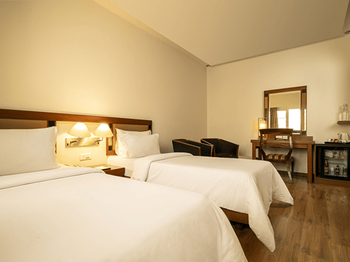 A warmly lit hotel room presents two single beds with white linens, a wooden desk with a mirror, and a mini-fridge at The Residency Karur.