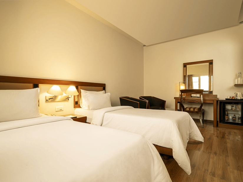 A warmly lit hotel room presents two single beds with white linens, a wooden desk with a mirror, and a mini-fridge at The Residency Karur.