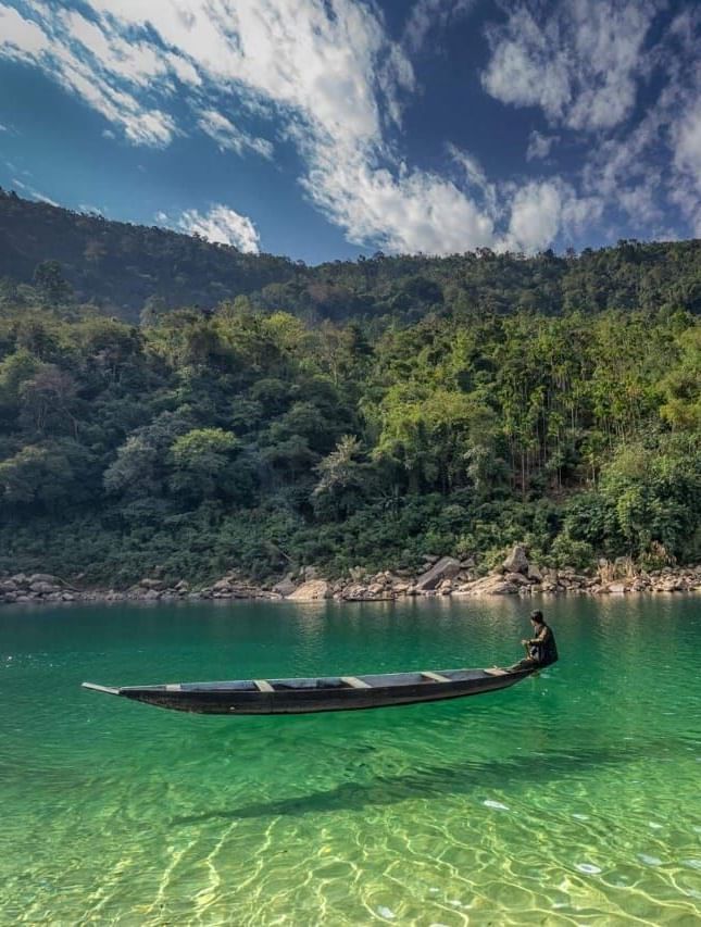 A serene river surrounded by lush forest with a lone wooden boat floating on the water - Polo Hotel Shillong