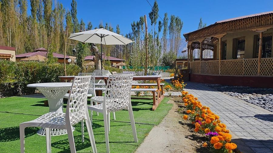 Outdoor dining area at White Sand Resort, Ladakh with white tables and chairs on a green lawn, surrounded by flowers and buildings.