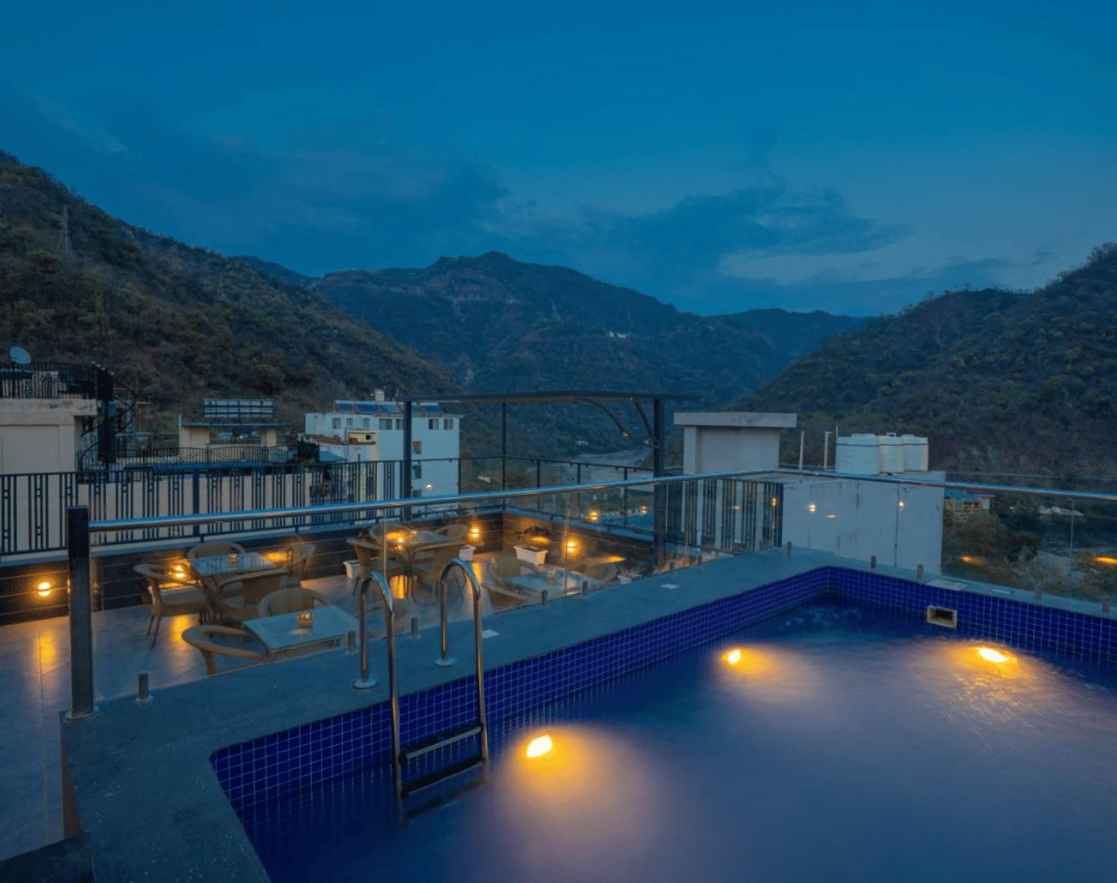 A rooftop cafe at Perfectstayz Premium Laxmi Heritage with an illuminated swimming pool and outdoor seating, overlooking mountains under a dark twilight sky.