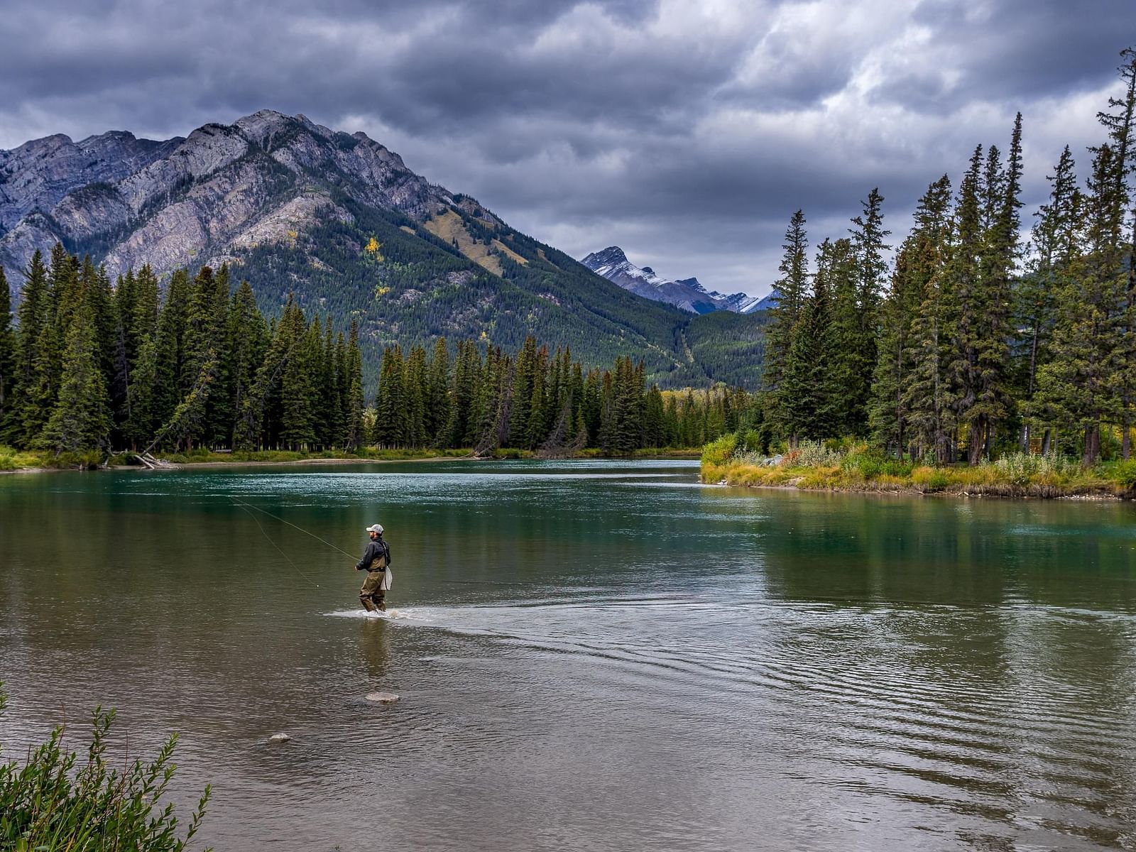 A lone person fishing in a river surrounded by greenery and a mountain.