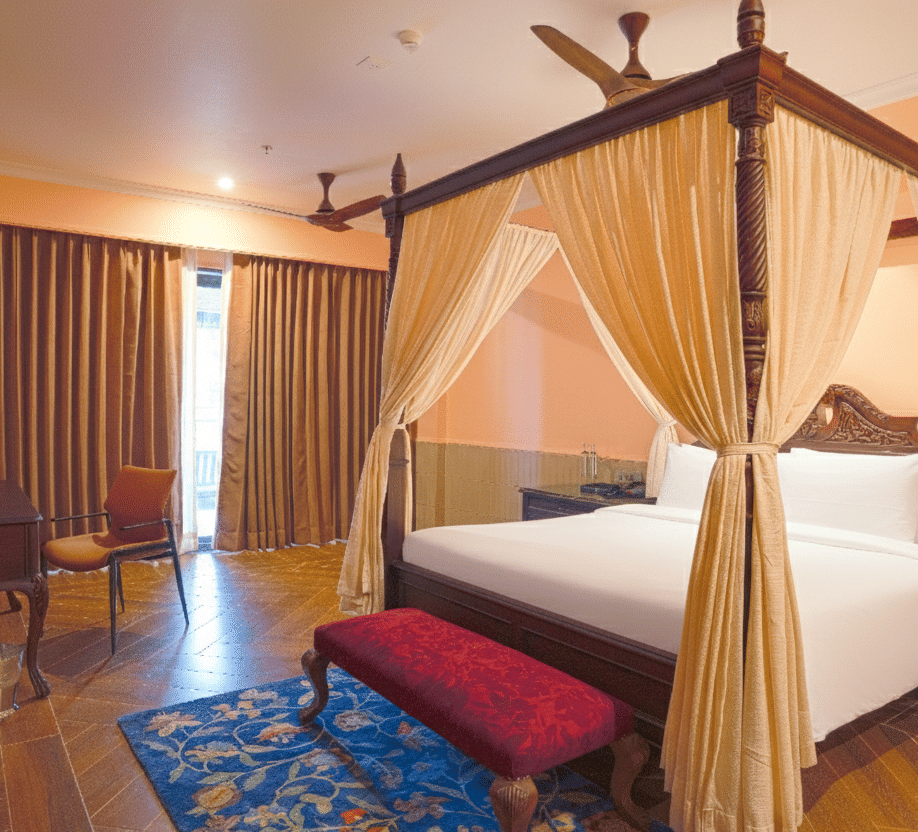 A bedroom with a four-poster bed draped in sheer curtains, wooden flooring, cushioned bench, sliding curtains and a small dining table set near the window at MAYFAIR Bay Resort, Paradeep.