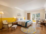 A bright hotel room with a queen bed featuring floral and teal bedding, a comfortable armchair with an ottoman, a wooden desk with a chair and a television, a floor lamp, and a balcony with a scenic view - Aloha On the Ganges.