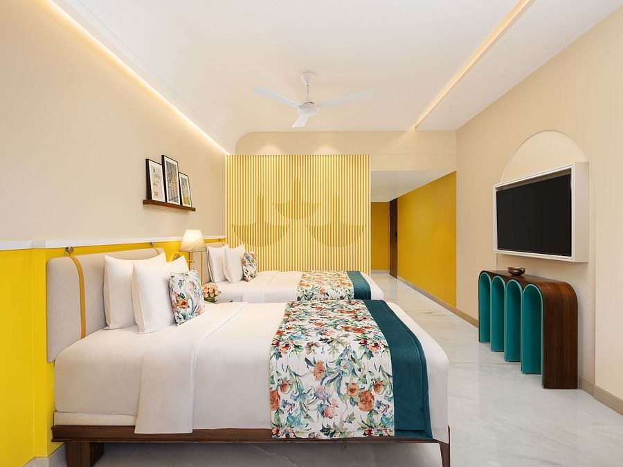 A hotel room with two twin beds featuring floral and teal throws, a stylish teal and brown cabinet with a television, a shelf with framed pictures, a ceiling fan, and yellow and white striped wall accents.