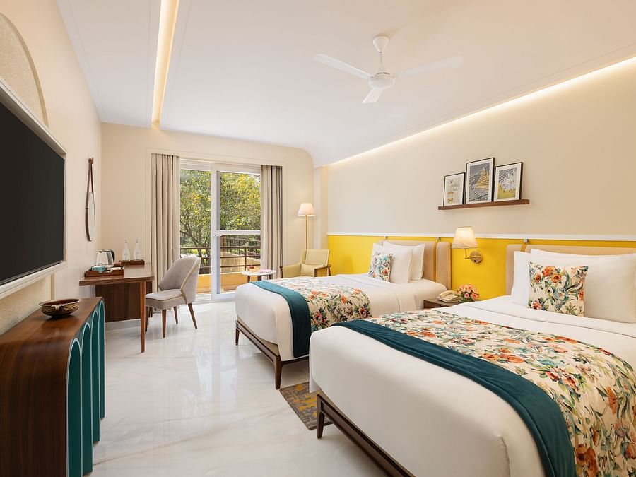 A bright hotel room with two twin beds featuring floral and teal accents, a wooden desk with a chair and a television, a stylish cabinet, a floor lamp, and a balcony with a view of trees - Aloha On the Ganges.