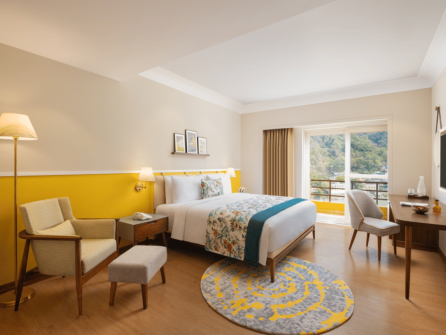 A bright hotel room with a queen bed featuring floral and teal bedding, a comfortable armchair with an ottoman, a wooden desk with a chair and a television, a floor lamp, and a balcony with a scenic view - Aloha On the Ganges.