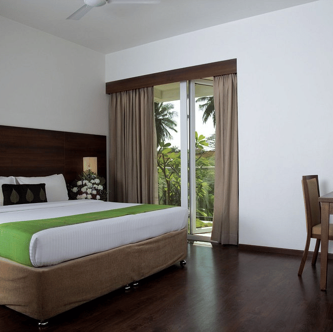 The Garden View Room with a king size bed dressed in crisp sheets and a bright  bed runner. To the right of the bed, there is a small wooden writing desk with a chair, and a large sliding glass door opening up to a balcony with a view of green trees at Tem