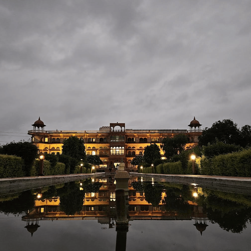 A well lit facade of Umaid Palace, a premiere resort near Bhangarh Fort, featuring stretched and multi storey building with lights on. The property is covered by trees and there is also a huge fountain in the front yard.