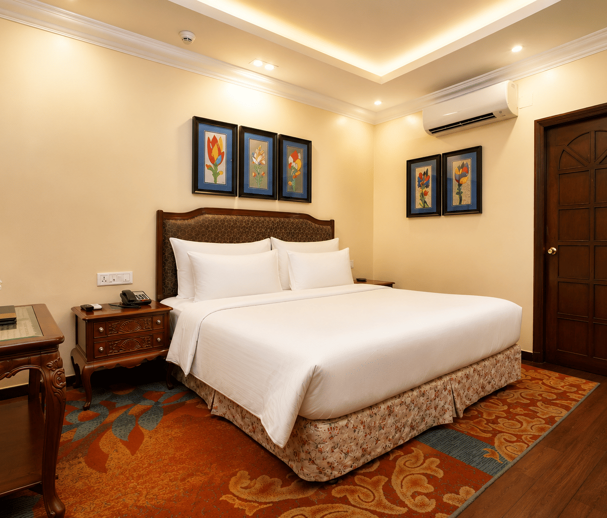 Executive room with a king bed, upholstered headboard, framed floral prints, bedside telephone, writing desk and a patterned carpet at MAYFAIR Garden, Rourkela.
