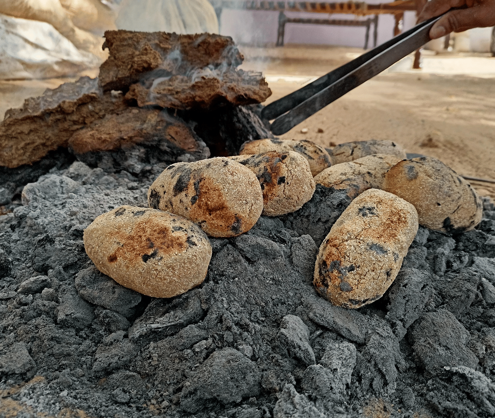 Charred potato-like dough balls (litti) are being cooked on a bed of hot grey ashes and glowing coals.