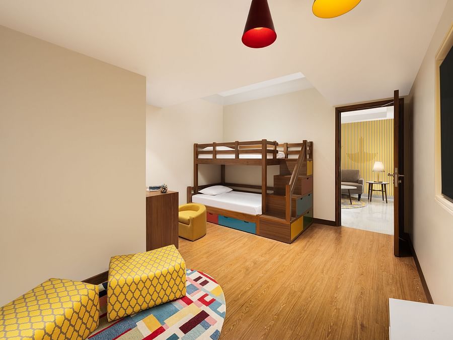 A children's bedroom with a wooden bunk bed featuring colourful storage drawers, a small yellow armchair, a wooden desk with toys, and a colourful patterned rug with yellow poufs, under modern pendant lights, with an open doorway leading to another room wi