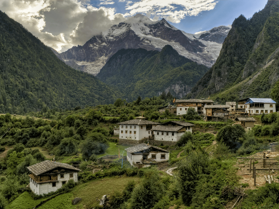 A scenic view of a remote mountain village with traditional white houses nestled in a lush green valley beneath jagged peaks.